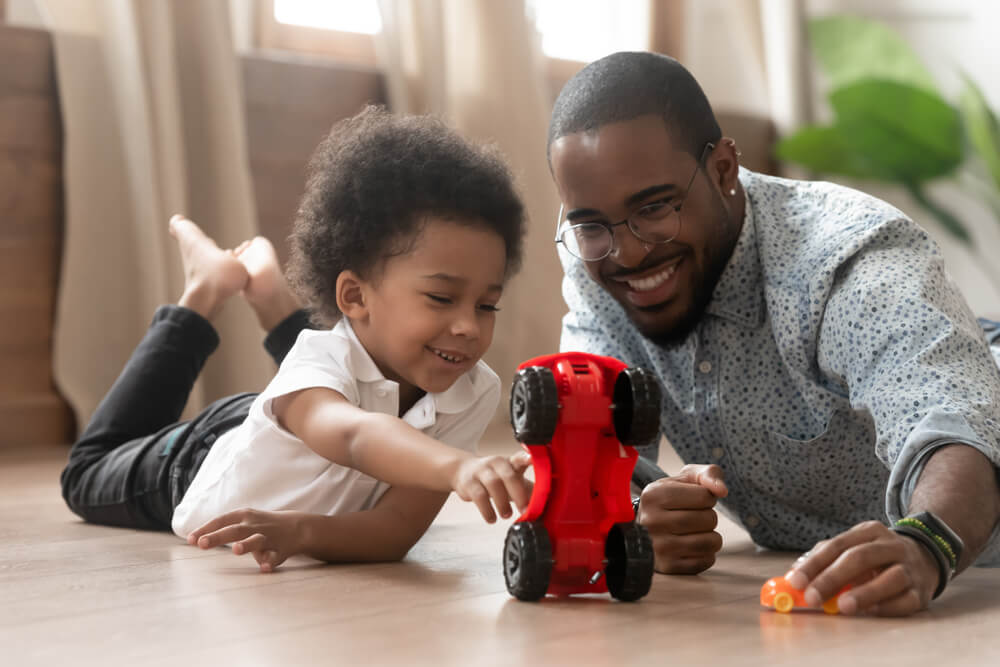 Two people lying on the floor playing with toy cars. One is reaching for a red toy car, and the other is holding an orange toy car. 