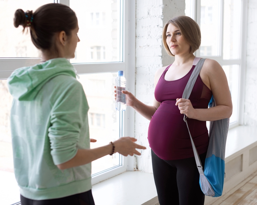 Two people stand near a large window. One wears a green hoodie; the other, visibly pregnant, holds a water bottle and carries a blue bag.