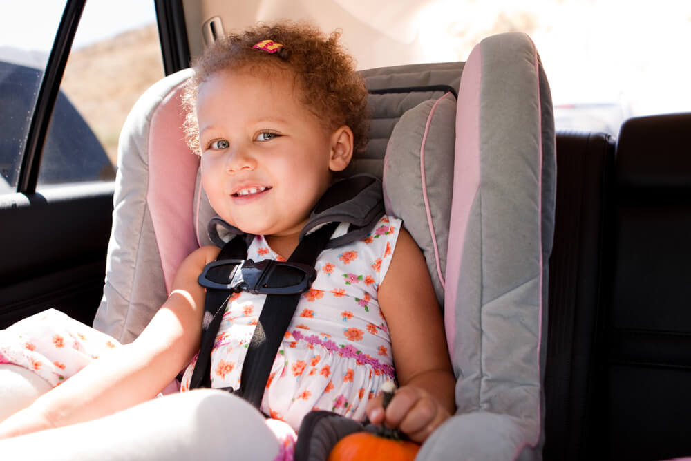 Un jeune enfant portant une robe blanche aux motifs floraux colorés est assis dans un siège d’auto gris montrant des accents roses à l’intérieur d’un véhicule.