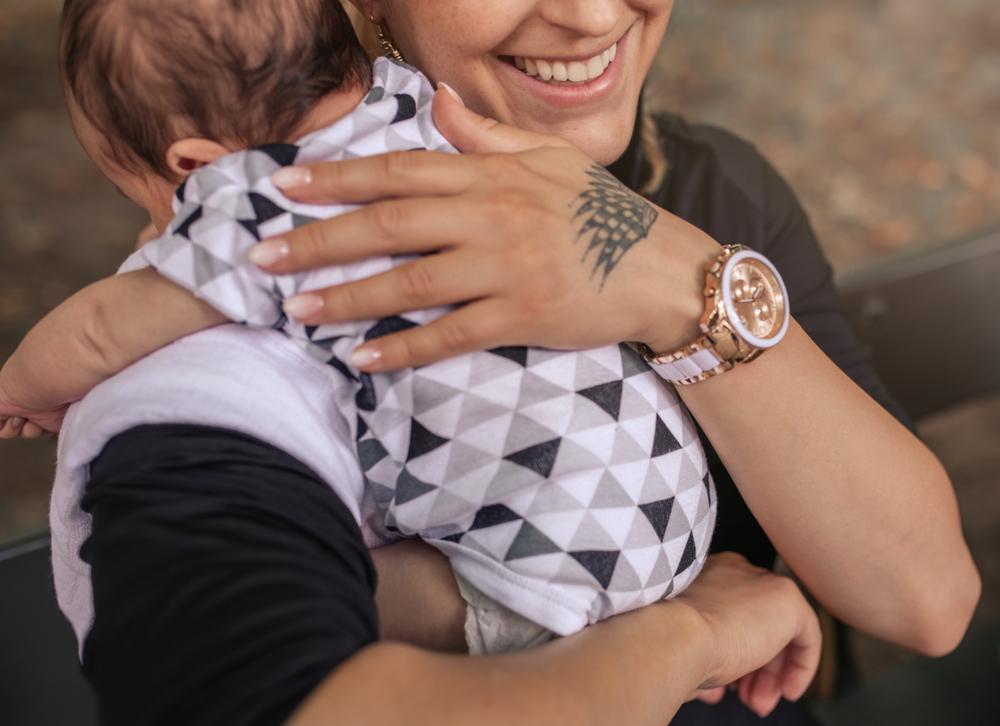 A person holds a baby dressed in a white and gray geometric outfit. The person has a hand tattoo, wears a wristwatch, and is partially smiling.