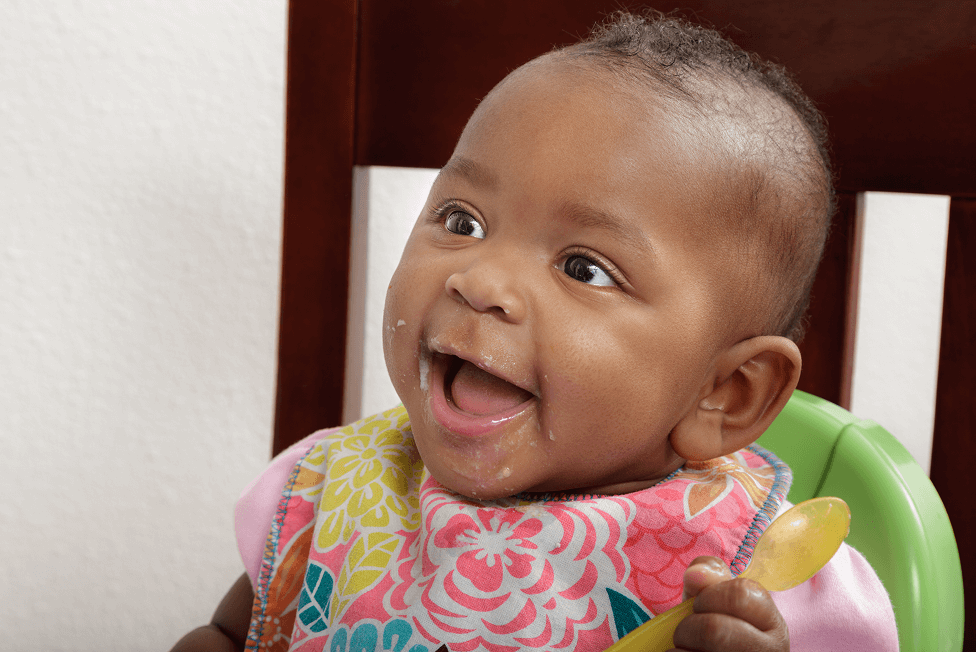 Baby in a high chair holding a yellow spoon, wearing a colorful bib.