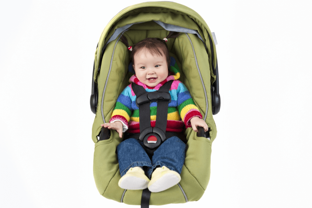 Un bébé portant un chandail rayé coloré, un jean bleu et des chaussures jaunes est assis dans un siège d’auto vert muni de sangles de sécurité noires.