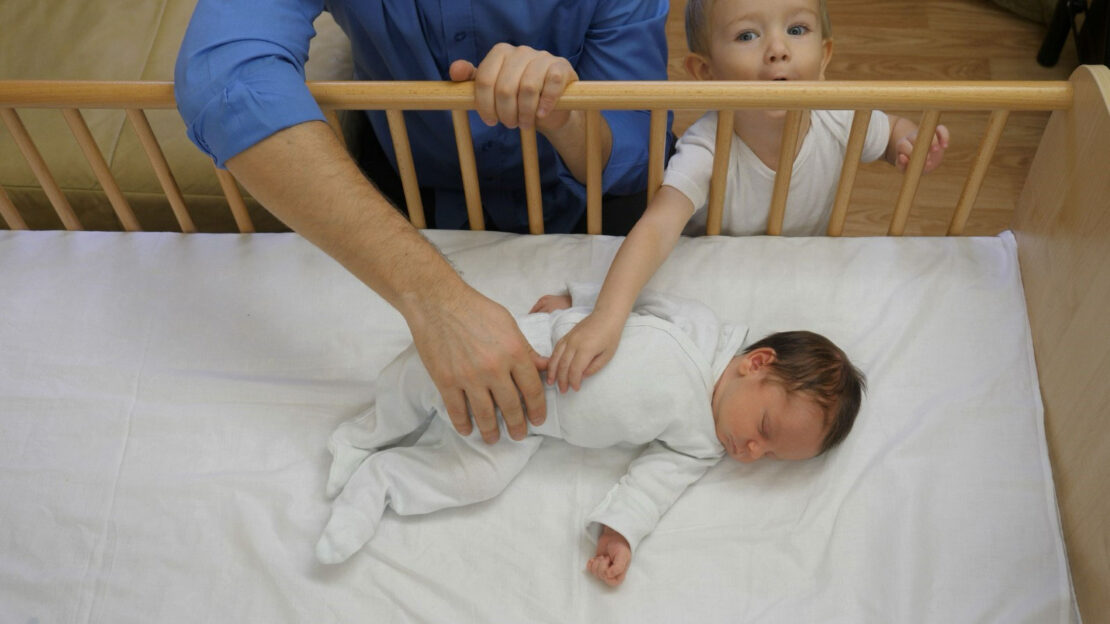 a baby in a white onesie sleeping on their back in a crib. An adult and a toddler are reaching in to gently touch them