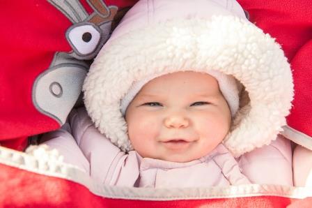 A baby dressed in a pink outfit with a white fur-lined hood is covered with a red blanket featuring an illustration of an animal, possibly a reindeer.