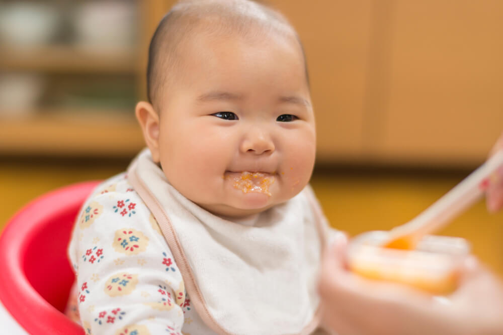 A baby sitting in a high chair wearing a bib is being fed by an adult hand holding a spoon. The background is blurred, drawing focus to the baby and the feeding moment