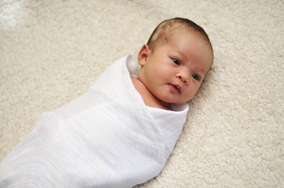 A baby wrapped in a white blanket lies on a soft, textured surface.