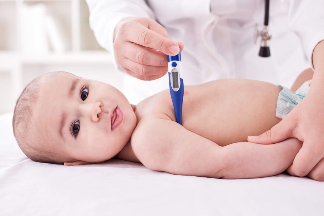 A baby lying on a white surface while a healthcare professional in a white coat with a stethoscope measures the baby's temperature using a blue and white digital thermometer under the baby's armpit.