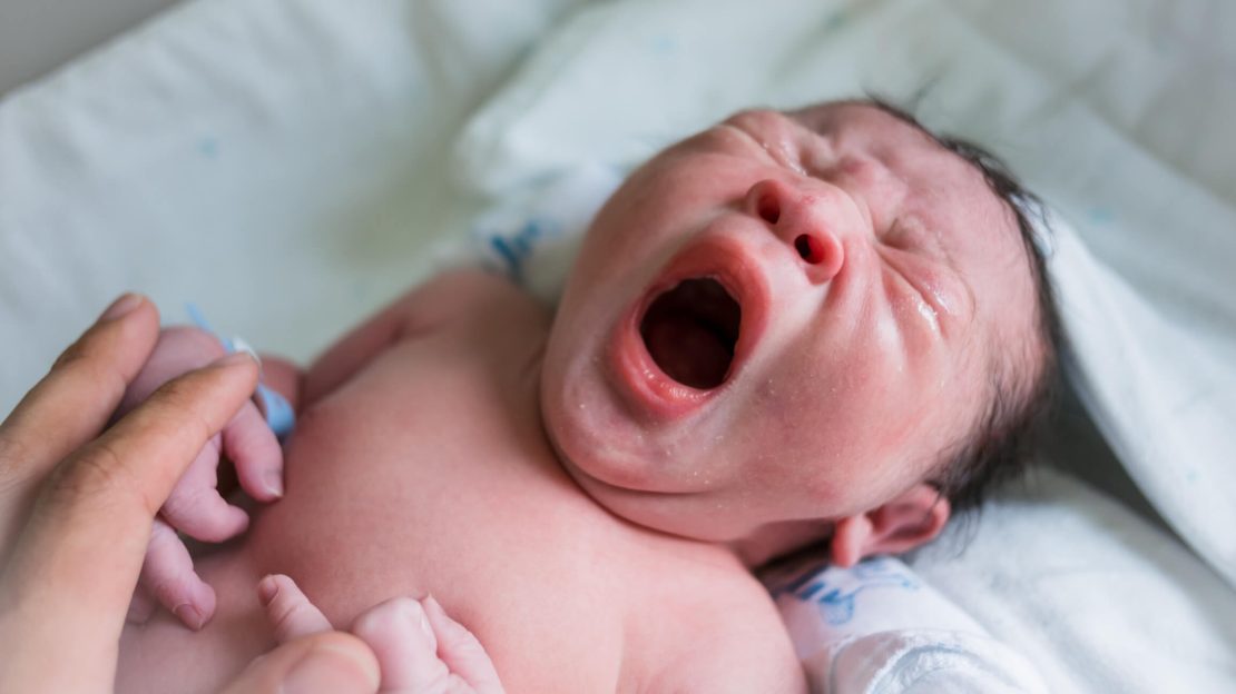 A newborn baby with dark hair lying on a white blanket, wrapped in a white cloth and an adult hand is gently holding the baby's left hand