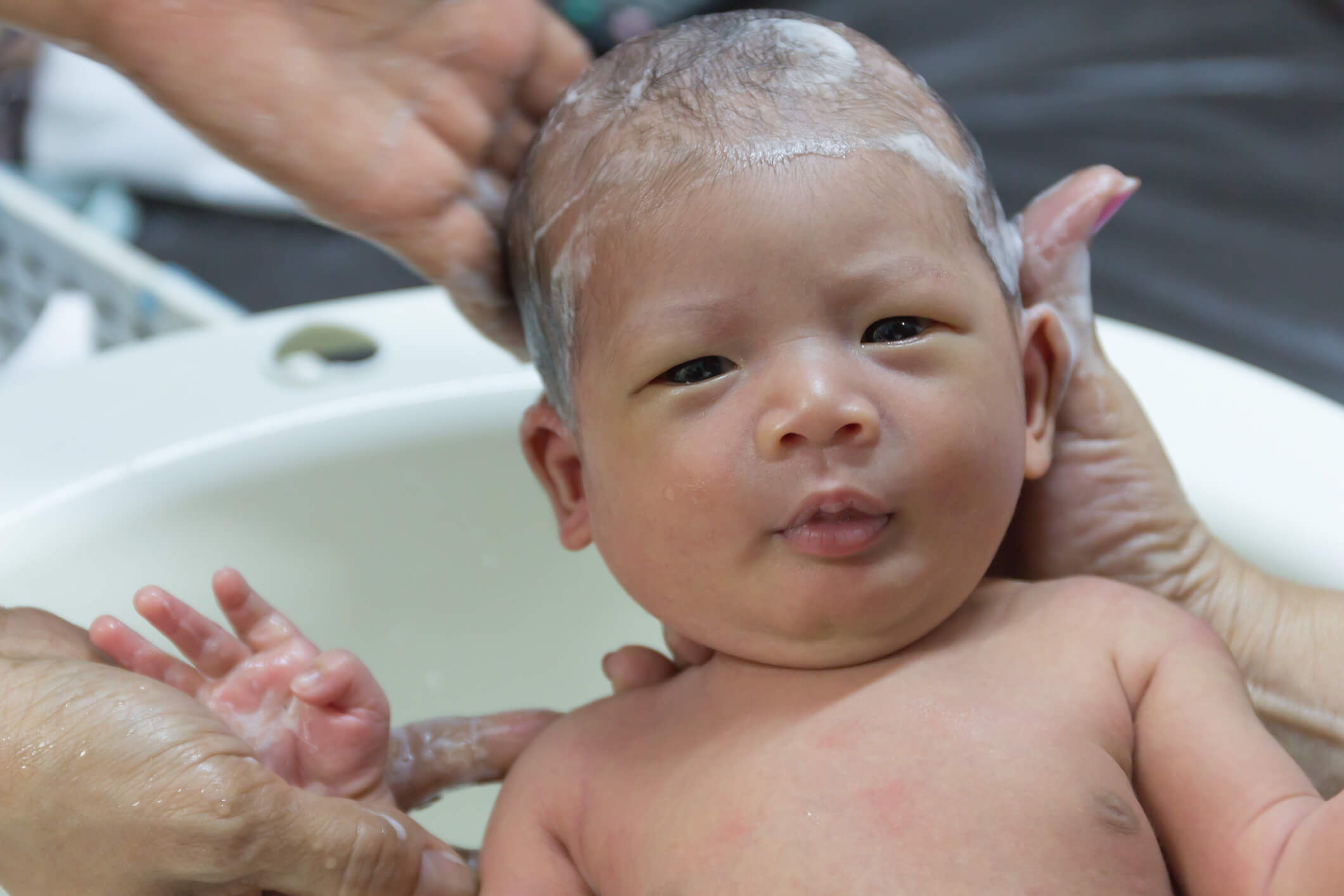 Un bébé prend un bain; des mains soutiennent et lavent sa tête à l’aide de savon ou de shampoing