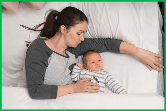 a parent and infant lying in bed together; the adult has their eyes closed and long hair in a ponytail. The infant has their eyes open and there are no sheets, comforters or pillows near the baby’s face