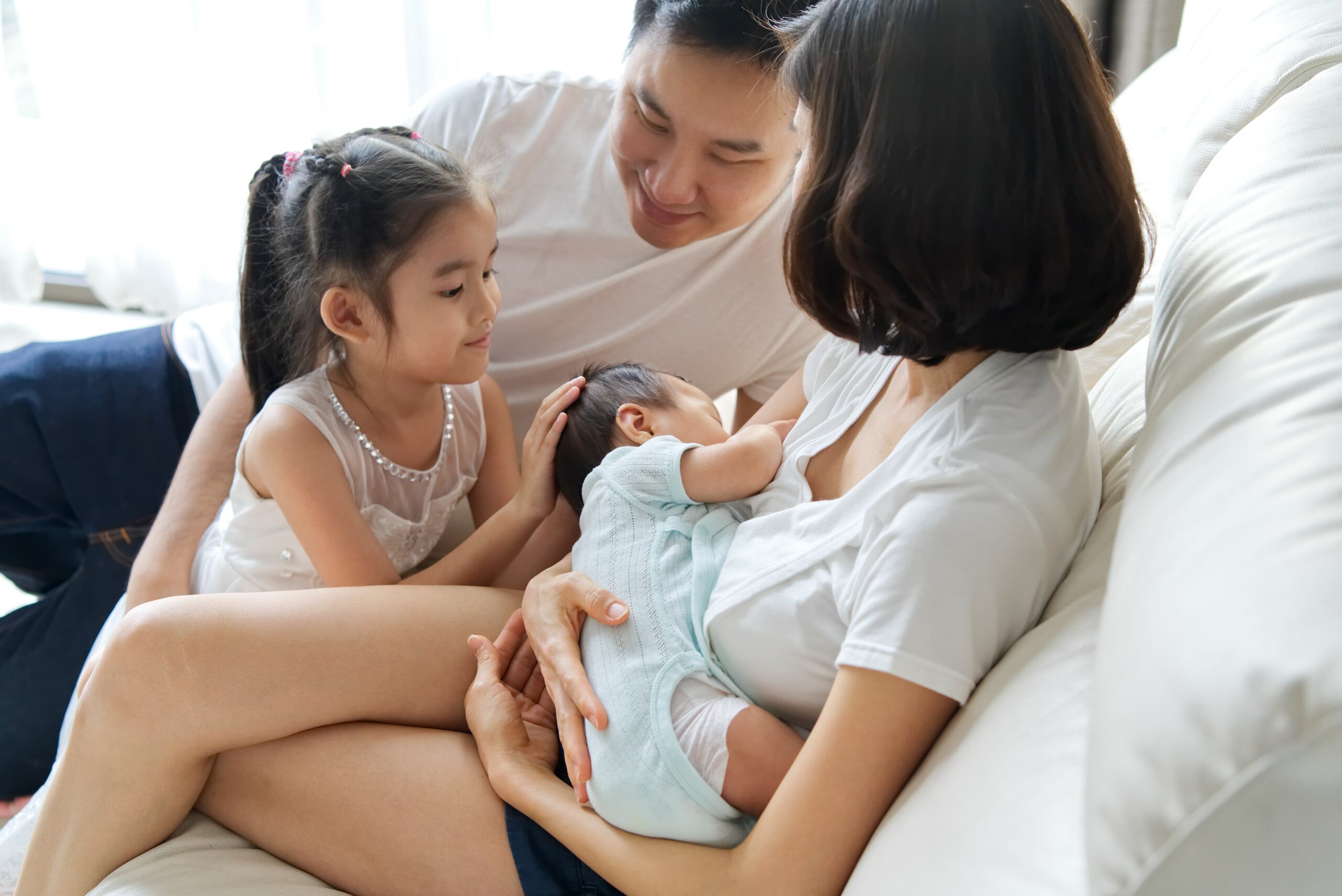 A family sits on a couch. One adult breastfeeds an infant while another child and adult sit nearby, looking at the baby. 