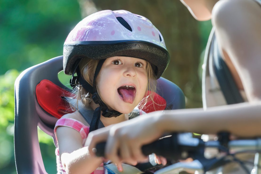 Un enfant portant un casque rose à pois est assis dans un siège de vélo, un adulte tenant le guidon. La scène se déroule en plein air, dans un lieu de verdure.