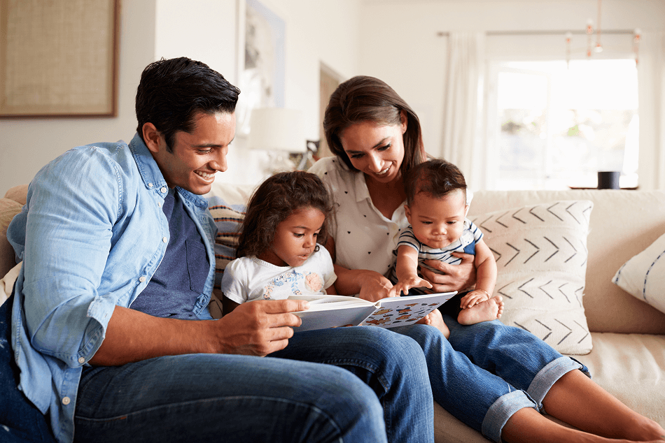 A family of four, with two young children and two adults, sitting on a sofa and reading a book together.