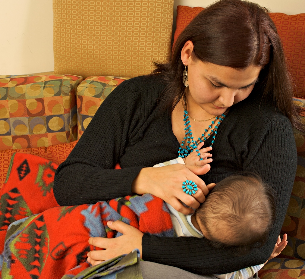Une personne allaite au sein un nourrisson enveloppé dans une couverture colorée. Elle porte une chemise noire et des bijoux de couleur turquoise, et est assise sur un canapé assorti de coussins à motifs.