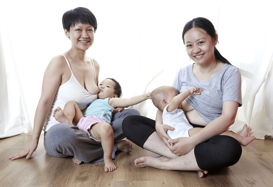 Two adults sit on the floor, each breastfeeding/chestfeeding a baby.