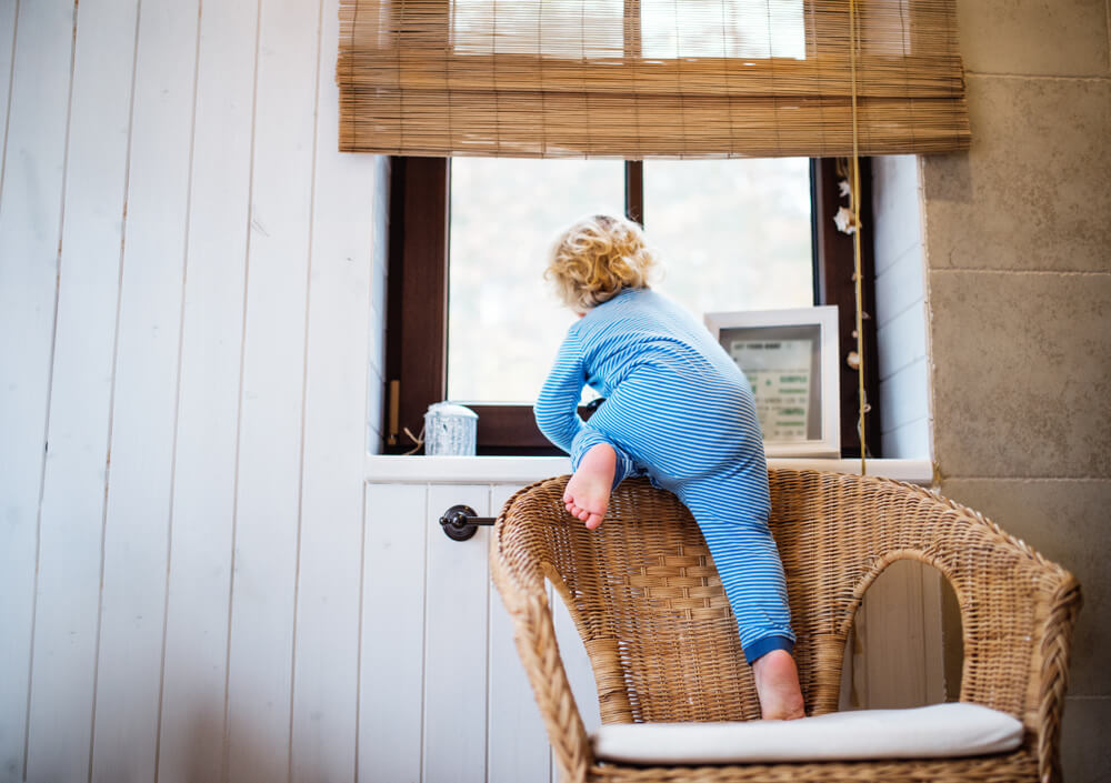 Un jeune enfant en pyjama bleu grimpe sur une chaise en osier pour regarder par une fenêtre munie de stores en bambou partiellement roulés. Une photo encadrée et un petit objet décoratif se trouvent sur l’appui de fenêtre. La chambre comprend des panneaux en bois blanc.