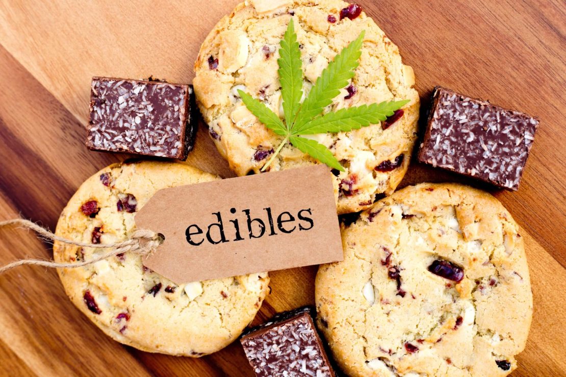 Trois biscuits et quatre carrés de chocolat disposés sur une surface en bois, avec une étiquette brune indiquant « Produits comestibles » et une feuille de cannabis placée sur un des biscuits.