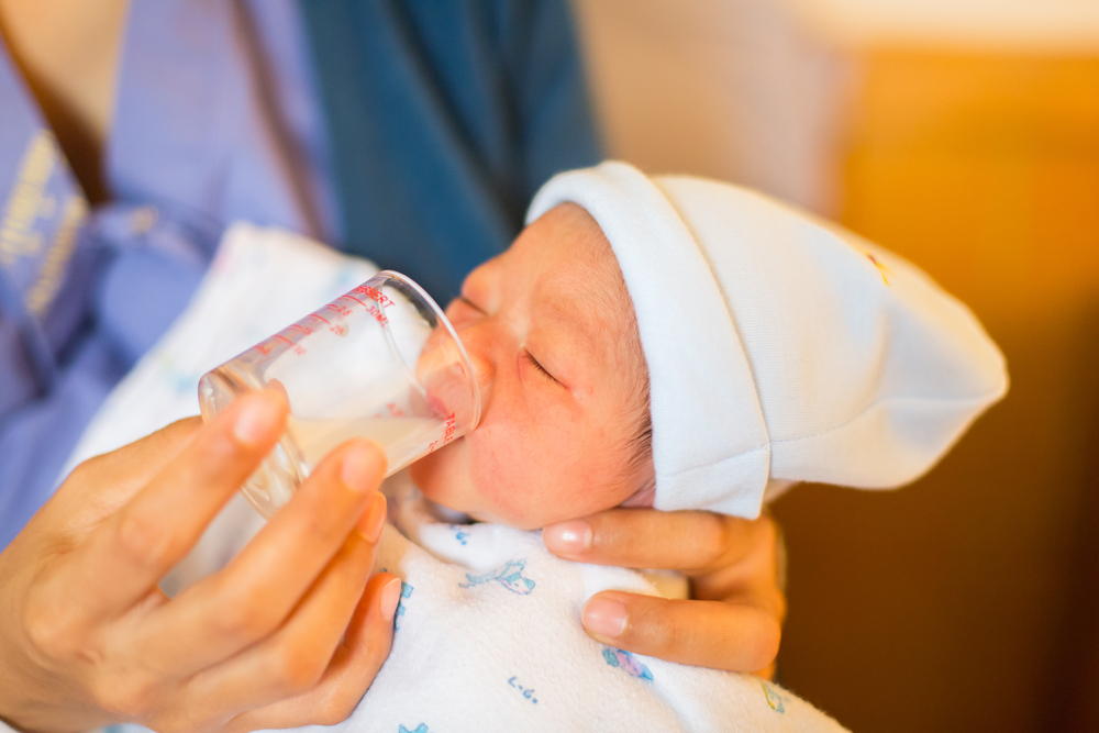 Un nouveau-né portant un chapeau blanc et une couverture boit du lait provenant d’une petite tasse à mesurer tenue par un adulte.