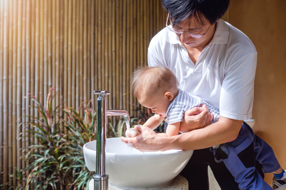 An adult holding a baby over a white sink, helping the baby wash hands under running water from a faucet. The adult is wearing a white shirt, and the baby is dressed in a blue striped outfit. The background features vertical bamboo sticks and some plants.