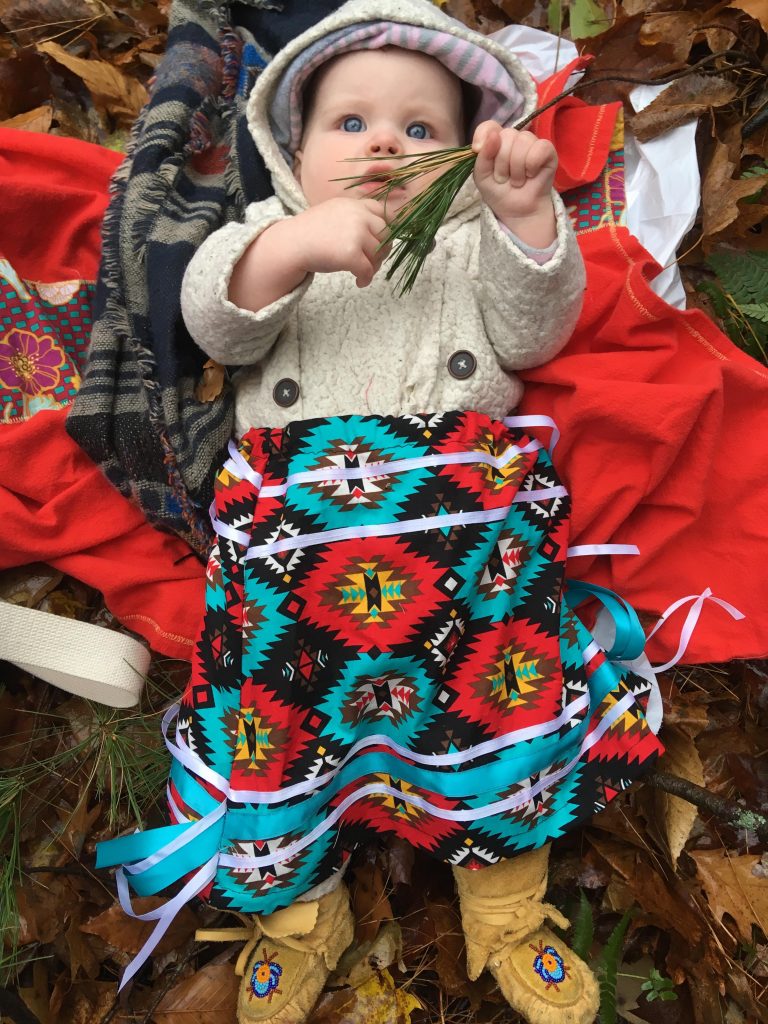 Enfant assis au sol entouré de feuilles et de couvertures colorées. L’enfant porte un manteau à capuchon, une jupe au motif géométrique vibrant et des mocassins, et tient un peu de produits verts dans une main.