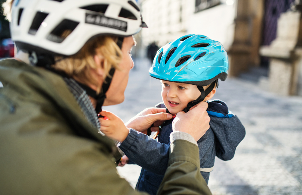Un adulte aide un enfant à mettre un casque bleu. Les deux portent un casque, et leurs visages sont flous. La scène se déroule à l’extérieur sur un pavé en cailloutis.
