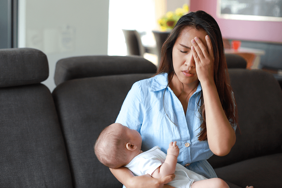 Une femme aux cheveux longs, assise sur un canapé, tient un bébé vêtu de blanc. Les yeux fermés, elle cache partiellement son visage de la main.