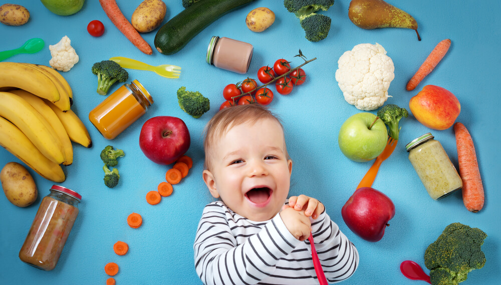 Bébé entouré de divers fruits, légumes et pots de nourriture pour bébé sur un fond bleu. Les articles comprennent des bananes, des pommes, des carottes, du brocoli, des tomates cerises, du chou-fleur, des pommes de terre, des zucchinis, des poires et des pots de purée d’aliments.