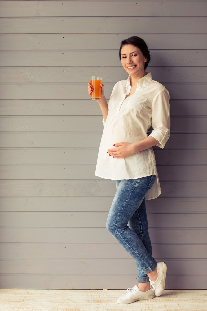 A smiling pregnant person holding a glass filled with orange liquid.