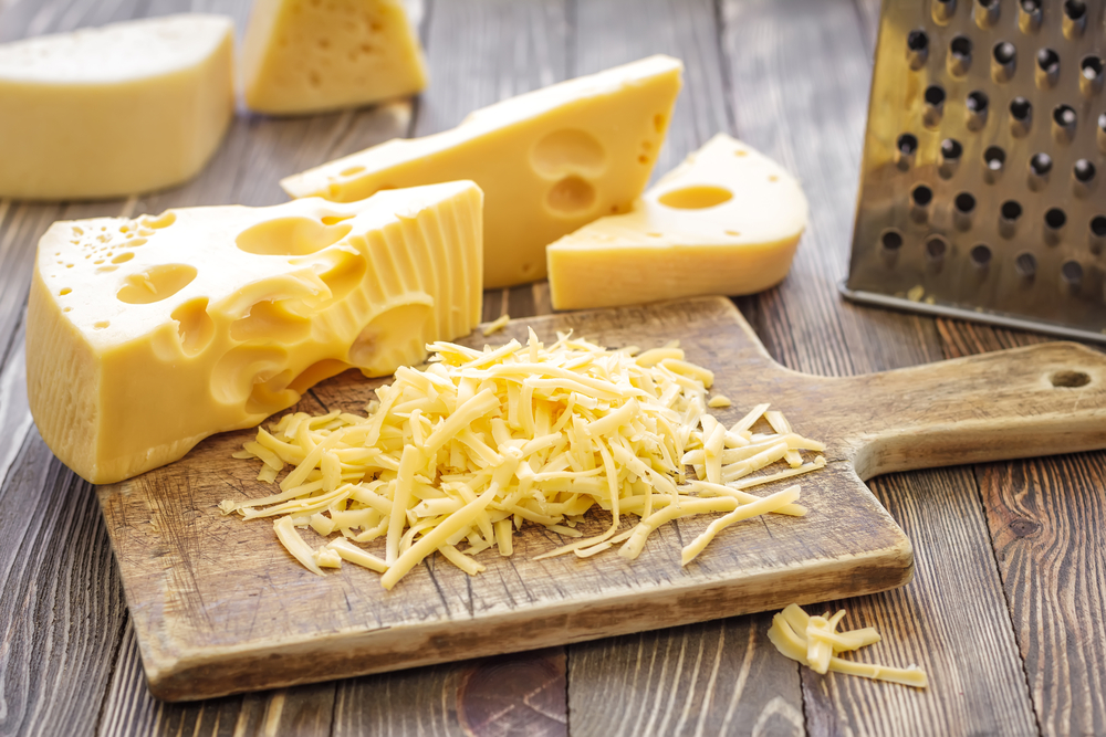 A pile of yellow shredded cheese and wedges of cheese on a wooden board.)