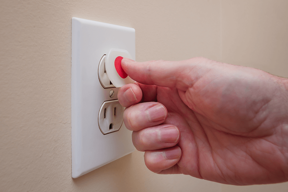 A close-up of a hand inserting a child safety plug with a red center into an electrical outlet on a beige wall