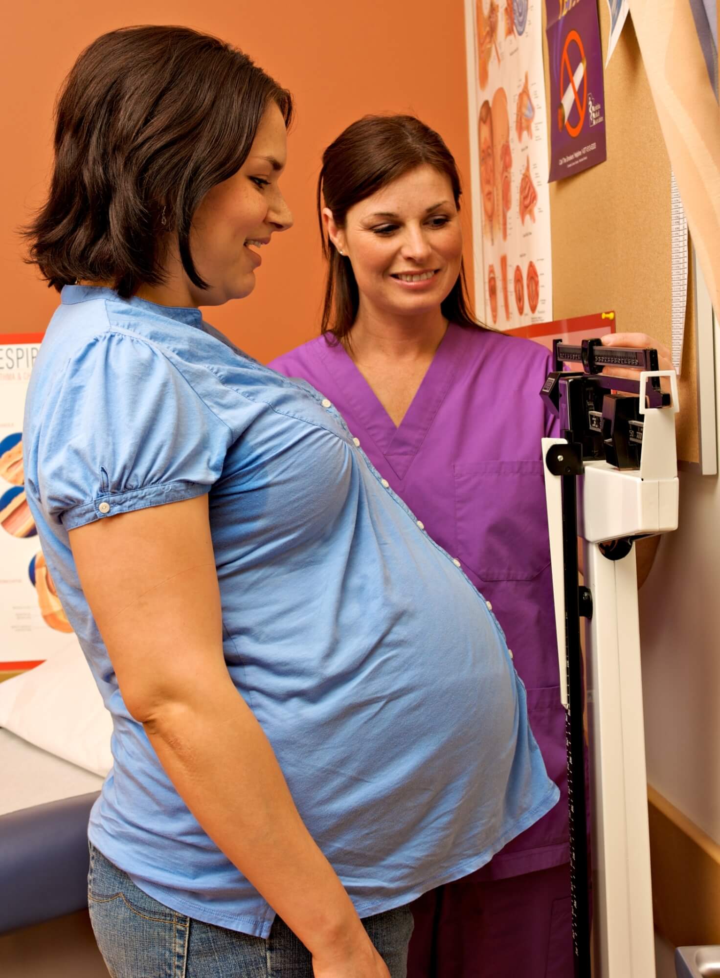 Personne enceinte vêtue d’une chemise bleue debout sur un pèse-personne, aidée d’un professionnel de la santé portant une blouse mauve. Des affiches et tableaux de médecine sont visibles sur le mur, en arrière-plan.