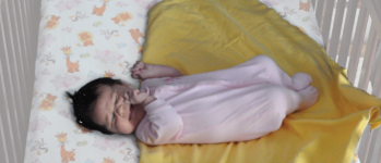a baby with dark hair in a light pink onesie on top of a yellow blanket with one corner folded