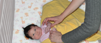 a baby with dark hair in a light pink onesie on top of a yellow blanket. An adult brings the baby’s hands to their chest