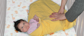 a baby with dark hair in a light pink onesie on top of a yellow blanket. An adult folds the bottom of the wrap up