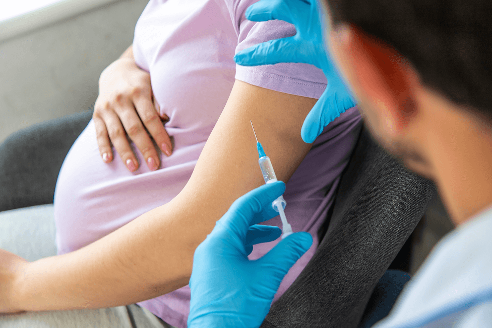 Pregnant person receiving an injection in their arm from a healthcare worker wearing blue gloves.