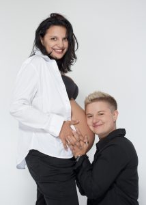 Smiling pregnant person standing while a partner kneels and holds their baby bump.