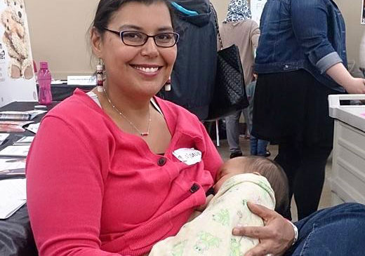 A person in a pink cardigan and glasses breastfeeds an infant wrapped in a light blanket. Other people and tables are visible in the background
