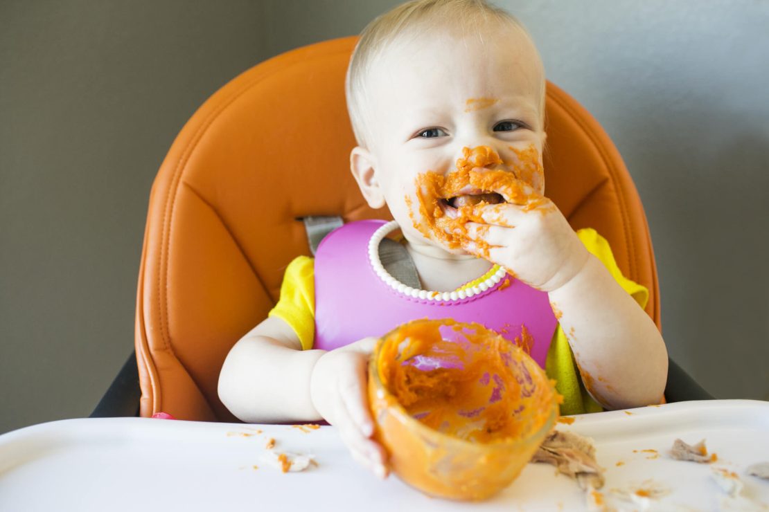 A child in an orange high chair wears a yellow shirt and pink bib, holding an empty bowl with food remnants. The tray and hands have traces of food