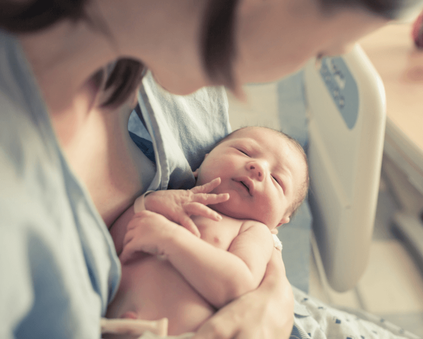 A person wearing a hospital gown holding a newborn baby in a hospital room. Medical equipment is visible in the background