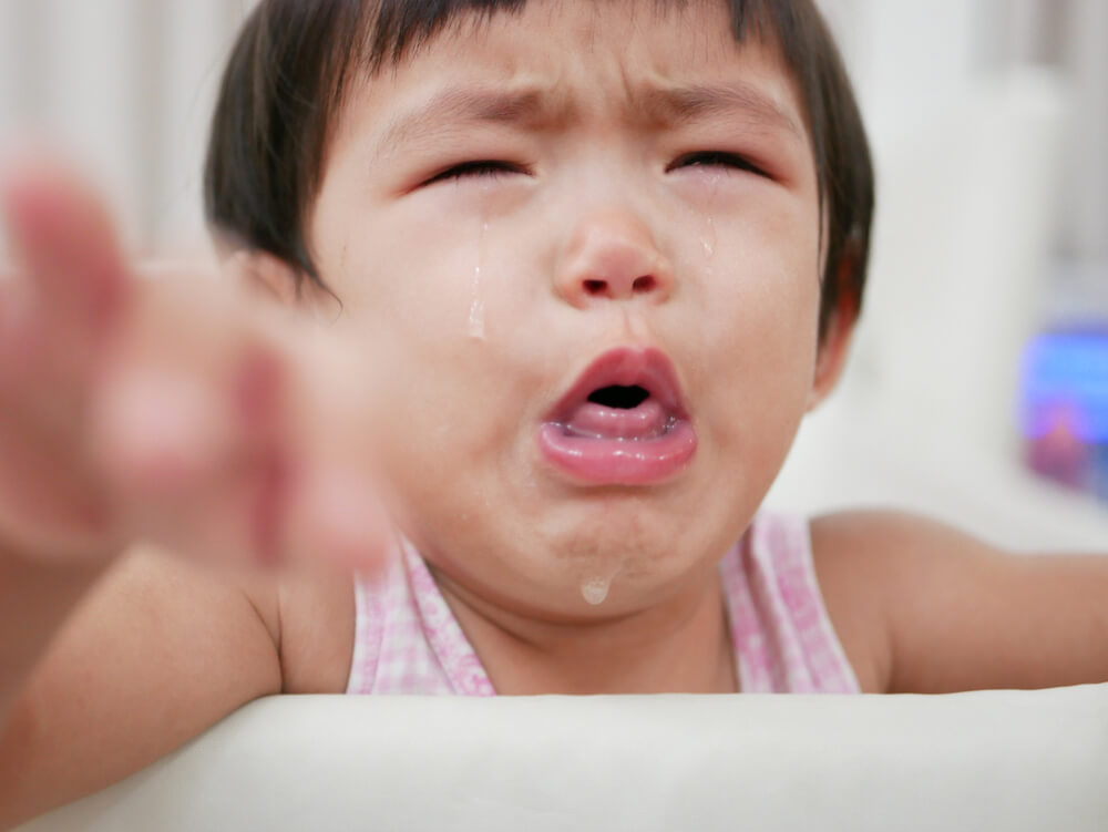 A young child with short dark hair wearing a sleeveless top, reaching out toward the camera. The background is indoors with indistinct objects