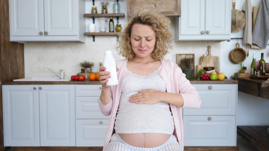 Personne enceinte assise dans une cuisine, tenant une bouteille blanche dans une main et reposant l’autre main sur son ventre. En arrière-plan, on voit des fruits, des légumes, des bouteilles et des ustensiles de cuisine.