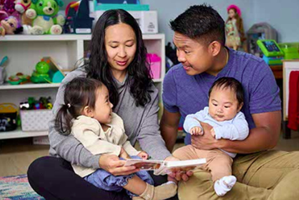 Family sitting on the floor reading a book and smiling.