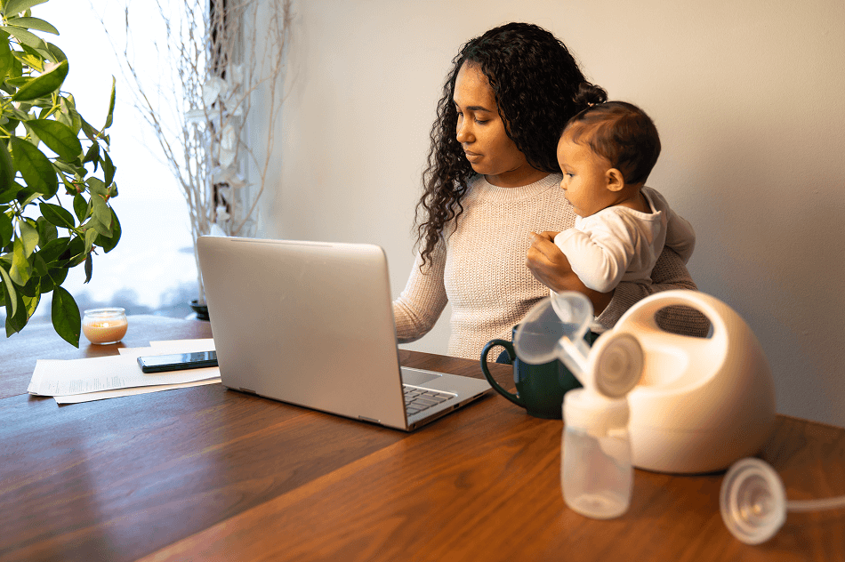 Une femme est assise à une table, travaillant sur un ordinateur portable tout en tenant son bébé. Un tire-lait et des biberons sont posés sur la table.