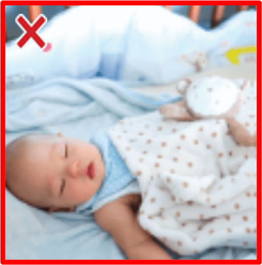 A baby asleep in a crib with a blanket and toy on top of them, and a bumper pad seen behind them. There is a red border around the image and an X in the top left corner.)