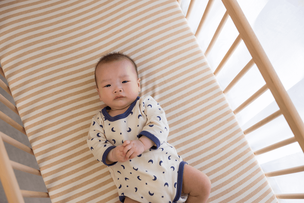 A baby in a white onesie with blue trim and crescent moon patterns lies in a wooden crib with a beige and white striped mattress.