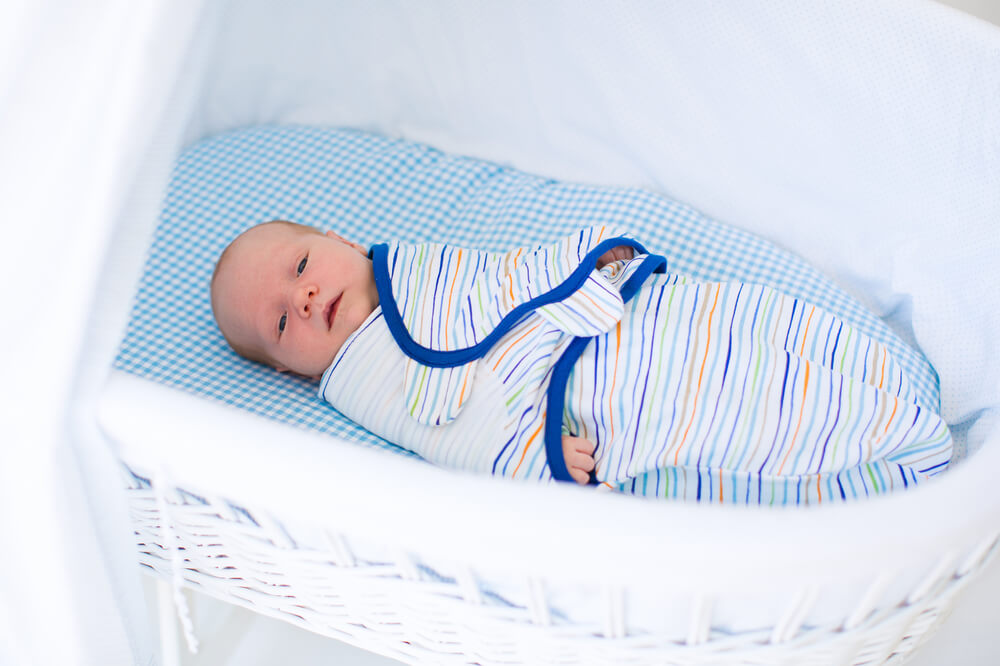 A baby wrapped in a striped swaddle with blue trim lies in a white wicker bassinet on a blue and white checkered mattress. 