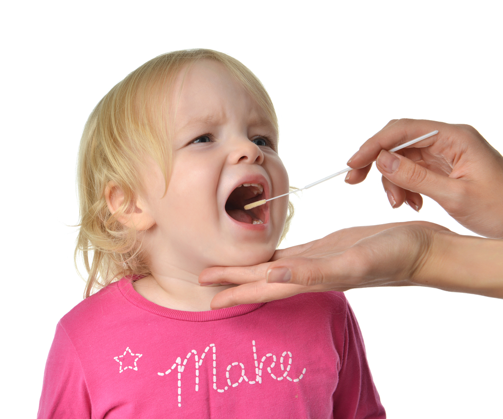 A young child with blonde hair wearing a pink shirt that says 'Make' in white dotted letters. An adult's hands are holding a cotton swab near the child's ear.