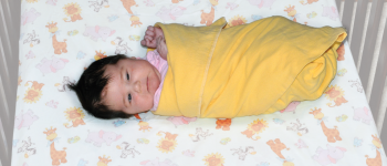 A baby is fully swaddled in a yellow blanket, lying on a crib mattress.  The baby's face is visible, and one hand peeks slightly out of the blanket.