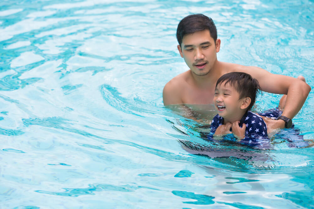 Two people in a swimming pool, with one holding the other, who appears to be a child wearing a blue and white star-patterned swimsuit. The water is clear and blue,