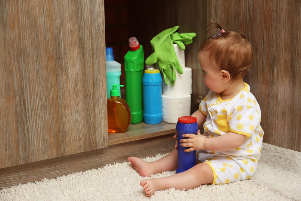 A young child sits on a carpeted floor in front of an open cabinet containing cleaning supplies, including bottles, green rubber gloves, and paper towels. The child is holding a blue bottle with a red cap. 